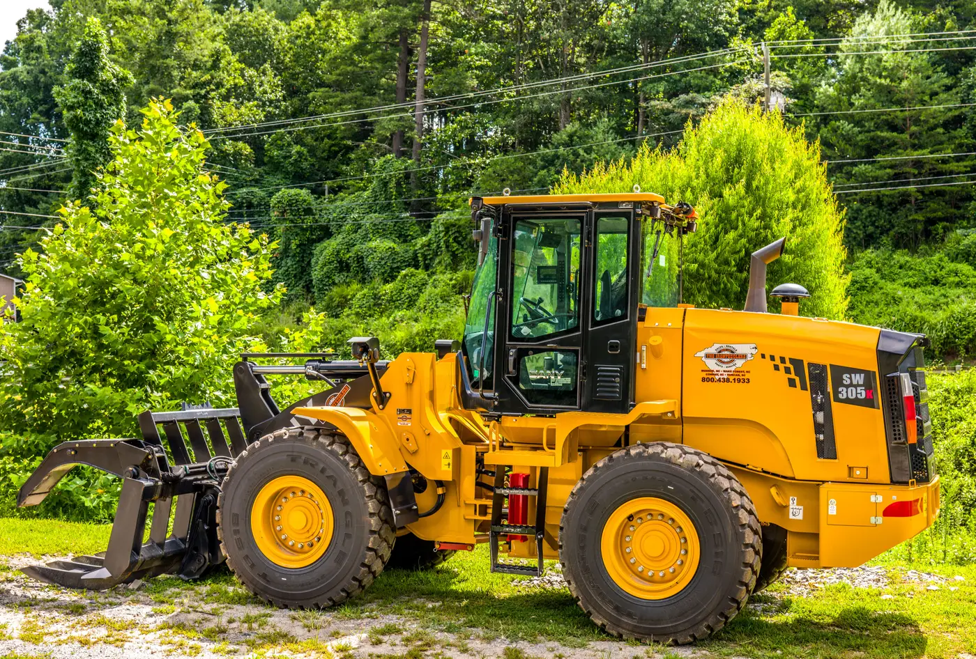 A yellow wheel loader with a bucket parked outdoors near trees in a timber yard setting.